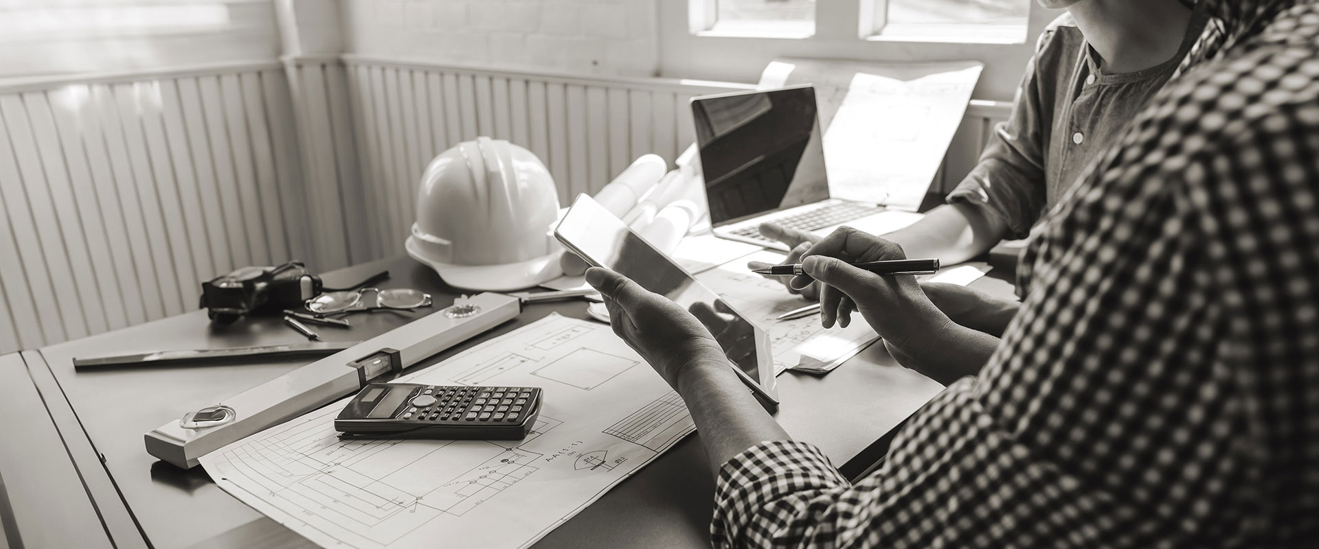 A construction site desktop with technical drawings, a laptop computer and a man holding a tablet.
